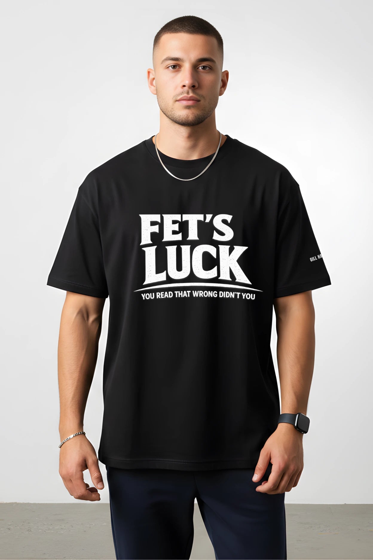 Man wearing black “FET’S LUCK; You Read That Wrong Didn’t You” graphic t-shirt by Gill Harry, featuring bold white distressed text and small sleeve logo, styled with navy joggers — witty streetwear for confident, humorous fashion lovers