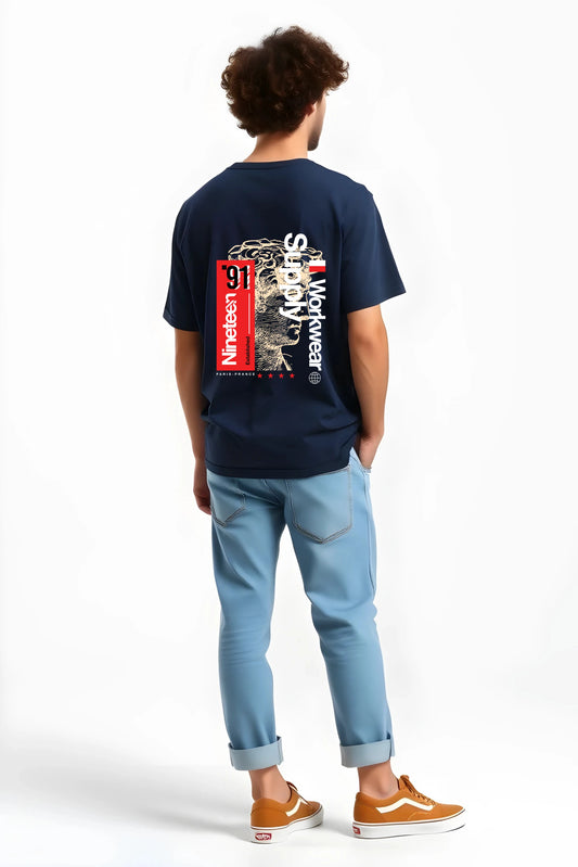 Back view of man wearing navy Nineteen ’91 Workwear Supply graphic t-shirt by Gill Harry, featuring bold red “Nineteen ’91” block and vintage-style face design, styled with light blue jeans and brown sneakers — minimalist streetwear with industrial edge