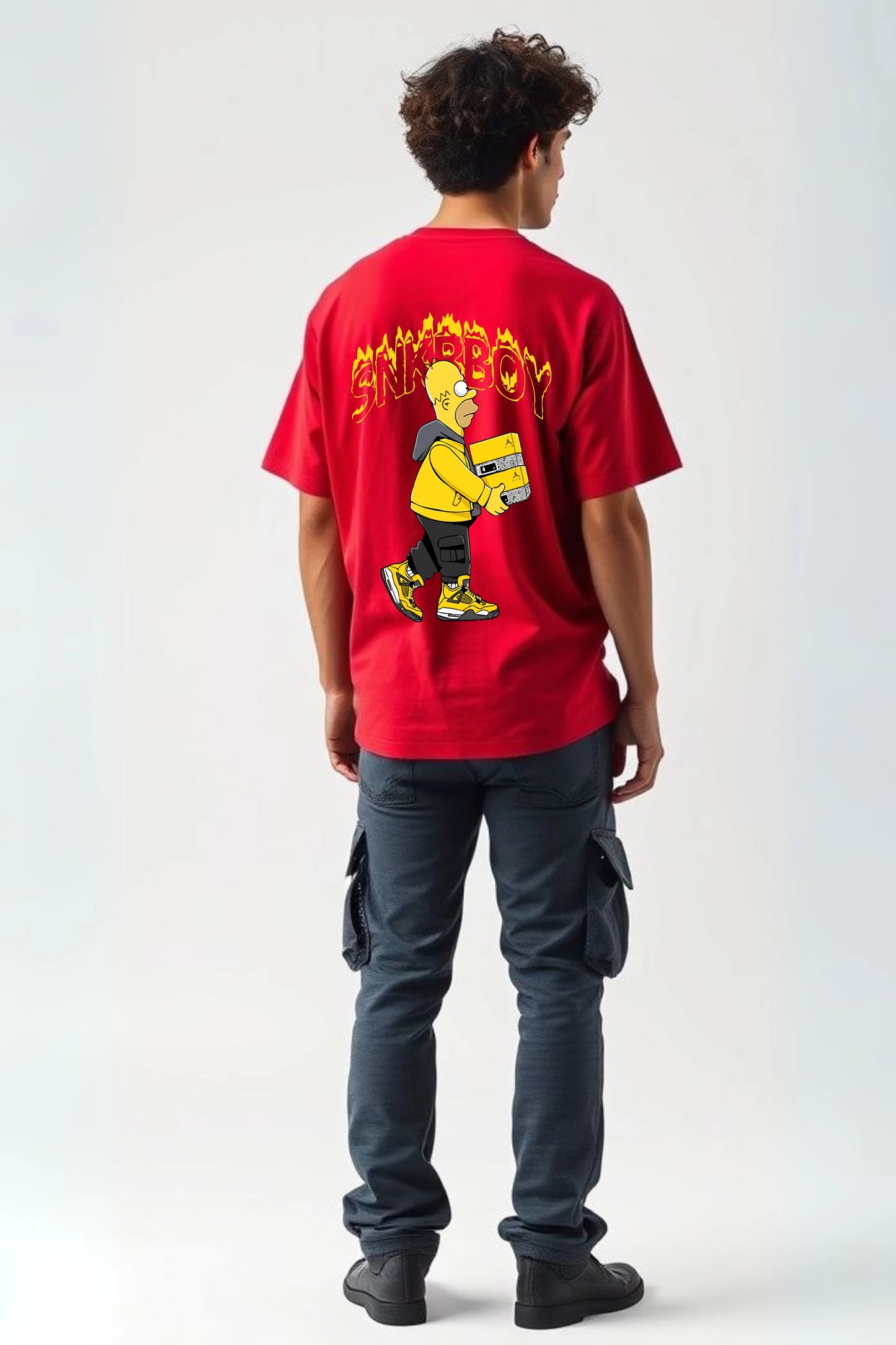 Back view of man wearing red SNKRBOY oversized graphic t-shirt by Gill Harry, featuring Homer Simpson as a sneakerhead holding a Jordan box, styled with dark cargo pants — bold streetwear for sneaker lovers and pop culture fans